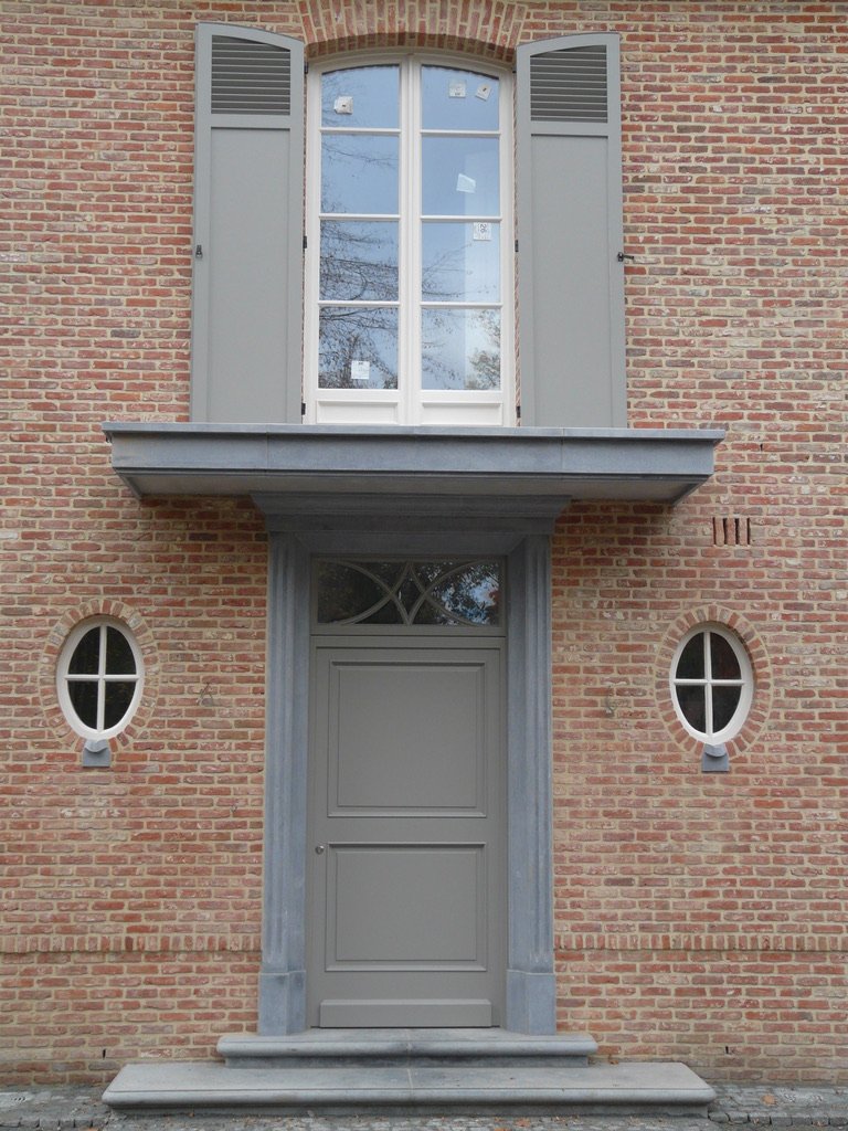 Statige grijze houten voordeur met bovenlicht en ovaal raampje, gecombineerd met luiken op de bovenverdieping.