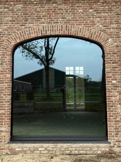 Groot houten boograam met strakke zwarte afwerking in een rustieke bakstenen gevel.