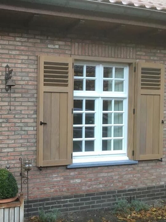 Wit houten raam met kruisverdeling, geflankeerd door rustieke eikenhouten luiken voor een warme uitstraling.