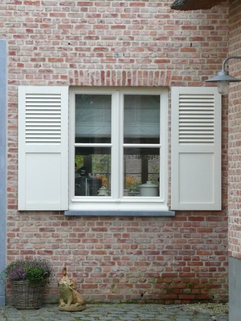 Landelijk wit houten raam met bijpassende witte klapluiken en lamellen, geplaatst in een bakstenen gevel.